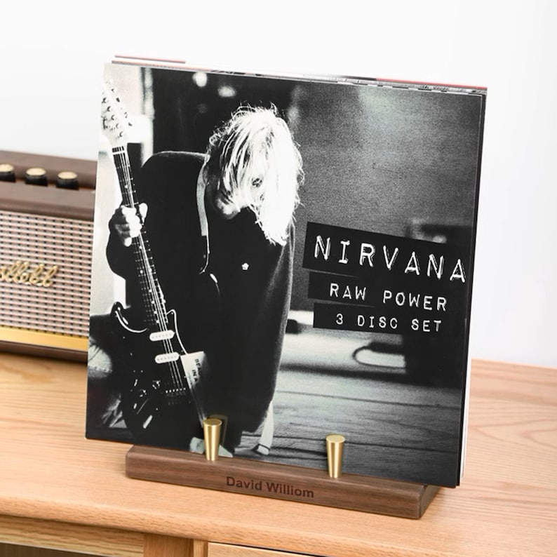 May include: A black and white image of the Nirvana "Raw Power" 3-disc set album cover, displayed on a wooden stand with brass pegs. The album cover features a photo of a person playing a guitar. The stand is engraved with the name "David William".