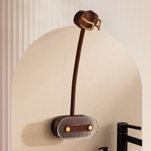 Pu&ograve; includere: Un portaspazzolino da parete con una base in legno marrone scuro e un frontale in acrilico trasparente e scanalato. Il supporto presenta un lungo e sottile stelo marrone e una sezione superiore con accenti metallici color oro.