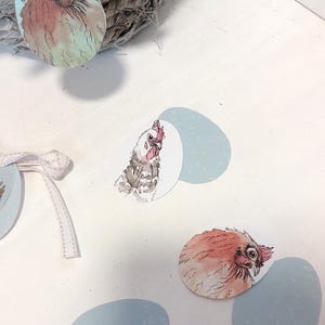 Könnte beinhalten: Aquarellillustrationen von Hühnern und Hähnen auf eiförmigen Ausschnitten. Die Bilder zeigen verschiedene Farben, darunter Rot, Orange und Braun. Einige Ausschnitte sind mit hellblauen Eiformen kombiniert. Im Hintergrund ein Weidenkorb mit blauem Band.