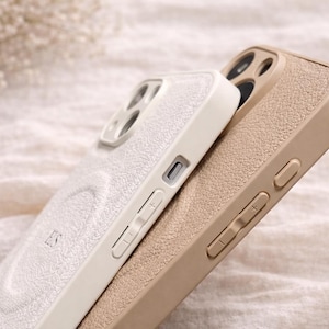 May include: Two phone cases, one off-white and one tan, are stacked together. The off-white case has a textured surface and the initials "ES" are visible. The tan case has a similar texture. Both cases have buttons and camera cutouts.