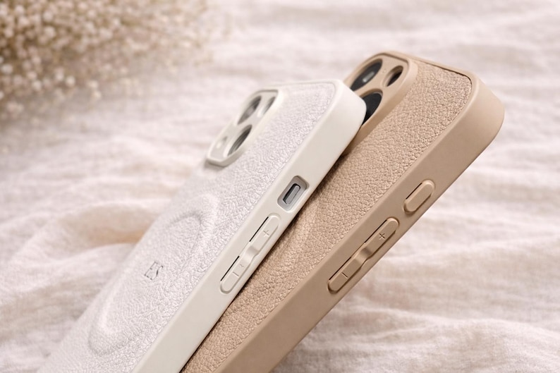 May include: Two phone cases, one off-white and one tan, are stacked together. The off-white case has a textured surface and the initials "ES" are visible. The tan case has a similar texture. Both cases have buttons and camera cutouts.