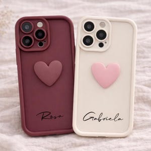 Może przedstawiać: Dwa etui na telefon: jedno bordowe, drugie kremowe. Każde ma serce i imię w skrypcie. Bordowe etui ma napis "Rosa", kremowe "Gabriela". Oba etui mają wycięcia na aparat.