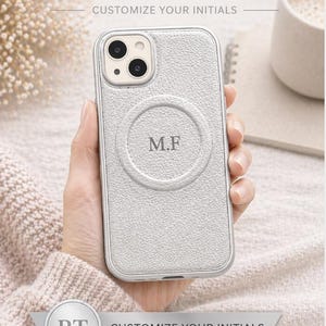 May include: A light gray leather phone case with the initials "M.F" embossed on the back. The case is personalized and features the text "CUSTOMIZE YOUR INITIALS". The case is designed for premium protection and has a genuine leather texture.