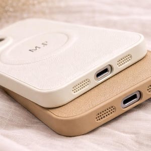 May include: Two leather phone cases, one cream-colored and the other tan, are stacked. The cream case has the initials "M.P." embossed on the back. Both cases have a textured finish and are designed with precision cutouts for the phone's ports and speakers.