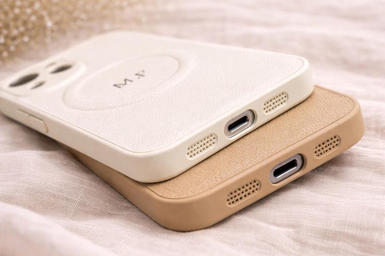 May include: Two leather phone cases, one cream-colored and the other tan, are stacked. The cream case has the initials "M.P." embossed on the back. Both cases have a textured finish and are designed with precision cutouts for the phone's ports and speakers.