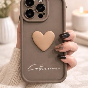 Może przedstawiać: Etui na telefon w kolorze szarobrązowym z wypukłym sercem i napisem "Catherine" w kursywie. Etui ma wycięcie na aparat z trzema obiektywami i lampą błyskową. Etui jest trzymane w dłoni.
