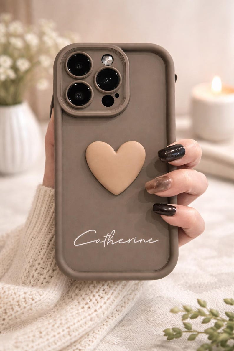 Może przedstawiać: Etui na telefon w kolorze szarobrązowym z wypukłym sercem i napisem "Catherine" w kursywie. Etui ma wycięcie na aparat z trzema obiektywami i lampą błyskową. Etui jest trzymane w dłoni.