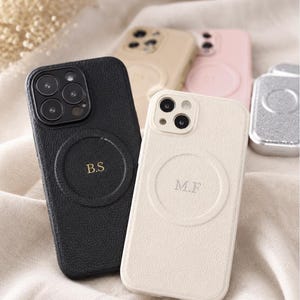 Puede incluir: Una colección de fundas de teléfono texturizadas con un anillo de carga magnético circular. La funda negra muestra las iniciales B.S en dorado, mientras que la funda crema presenta las iniciales M.F. Se ven fundas adicionales en beige, rosa y plata.