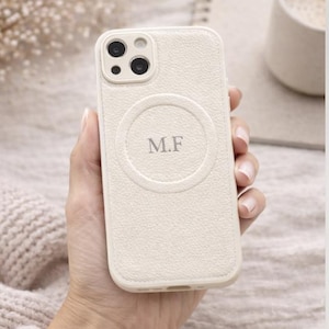 May include: A cream-coloured phone case with a textured finish, featuring a circular design with the initials "M.F." The case has a camera cutout. The text "CUSTOMIZE YOUR INITIALS" is at the bottom. The phone case is held in a hand.