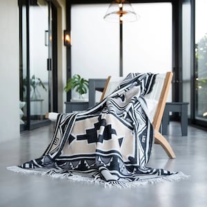 May include: A black, white, and beige patterned throw blanket with fringe is draped over a wooden chair. The chair is in a modern, minimalist room with large windows.