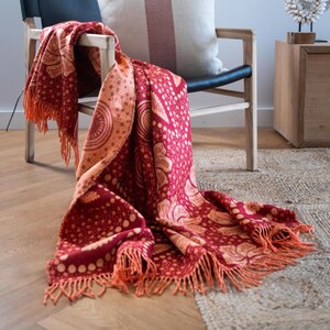 May include: A red and peach-colored throw blanket with a detailed pattern and fringe. The blanket is draped over a wooden chair, showcasing its design and texture. The throw features a repeating circular and dotted pattern.