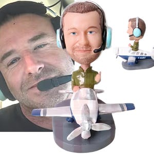 May include: A custom bobblehead figurine depicting a person wearing a green shirt and a headset with light blue ear cups, seated in a small white and blue airplane with a propeller. The figurine is mounted on a gray base, with a side view included.