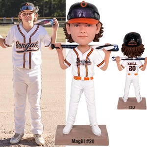 Op de afbeelding: Een op maat gemaakte bobblehead-figuur van een honkbalspeler in een wit uniform met oranje en zwarte biezen, het woord Bengals en de naam Magill #20. Het figuur draagt een zwarte helm, houdt een honkbalknuppel vast en staat op een voetstuk.