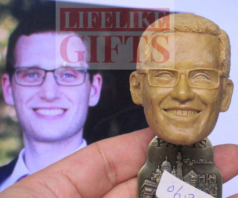 Pu&ograve; includere: Una statuetta bobblehead sorridente di un uomo con gli occhiali, con un viso e capelli dettagliati. La base della statuetta ha un design di paesaggio urbano e la parola "PARIS". Sullo sfondo c'&egrave; una foto di un uomo con gli occhiali.