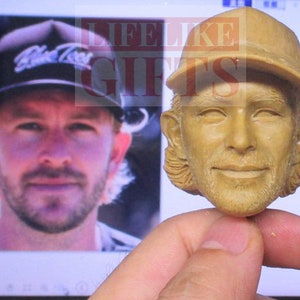 Pu&ograve; includere: Un busto scolpito color marrone chiaro di un uomo con un berretto da baseball. Il busto &egrave; tenuto davanti a uno schermo di computer che mostra una foto dello stesso uomo con un berretto da baseball. Il berretto nella foto dice "Blue Tees".