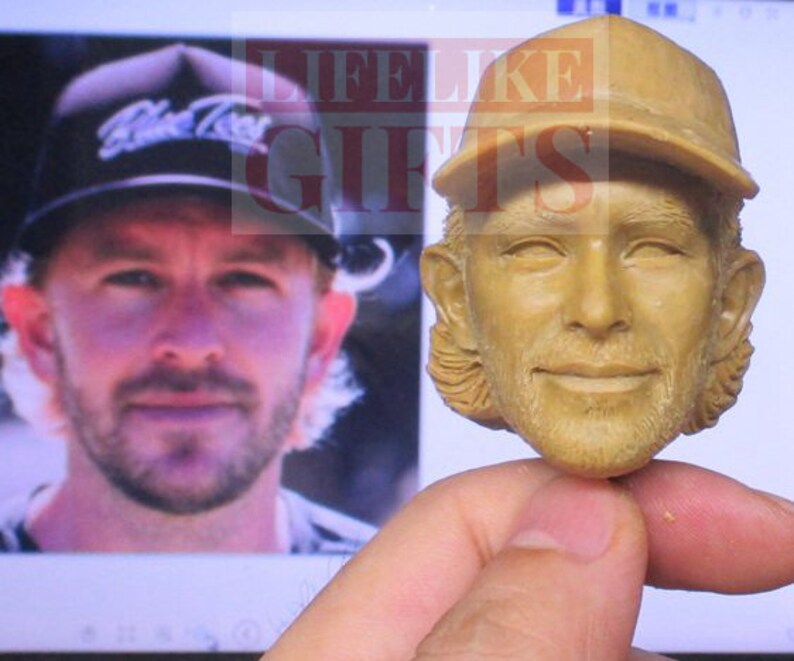Pu&ograve; includere: Un busto scolpito color marrone chiaro di un uomo con un berretto da baseball. Il busto &egrave; tenuto davanti a uno schermo di computer che mostra una foto dello stesso uomo con un berretto da baseball. Il berretto nella foto dice "Blue Tees".
