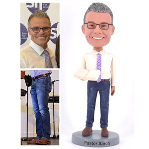 May include: A custom bobblehead figurine of a man with gray hair, wearing a white shirt, a purple tie, and blue jeans. He is holding a book in his right hand. The base of the figurine is gray and has the text "Pastor Aaron" printed on it.