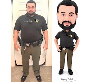 May include: A custom bobblehead figurine depicting a person with a dark beard wearing a black polo shirt, olive green trousers, and a black utility belt with a badge. The figurine stands on a white circular base with the text Deputy Colon printed.
