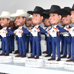 May include: A group of 15 bobbleheads, all wearing blue suits and cowboy hats, are lined up on a shelf. Each bobblehead has a different face and is holding a white shirt in front of them. The bobbleheads are all facing forward and have a white background.