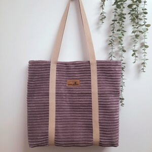Könnte beinhalten: Eine lilafarbene Cord-Tragetasche mit cremefarbenen Henkeln. Die Tasche hat einen kleinen braunen Leder-Patch mit dem Text "ARICA DESIGNS" darauf.