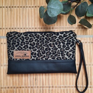Könnte beinhalten: Eine schwarze und goldene Clutch mit Leopardenmuster, schwarzem Lederboden und schwarzem Riemen. Die Clutch hat ein braunes Lederetikett mit der Aufschrift "Handmade Handbags Shop" und "Annika Biergans London".