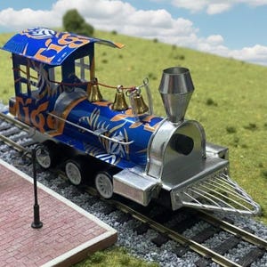 Könnte beinhalten: Ein Miniaturmodell eines Zuges mit blauem und orangefarbenem Design, einem silbernen Schornstein und Glocken. Der Zug fährt auf einer Strecke neben einem Miniatur-Bahnhof mit dem Namen "HENRIET".