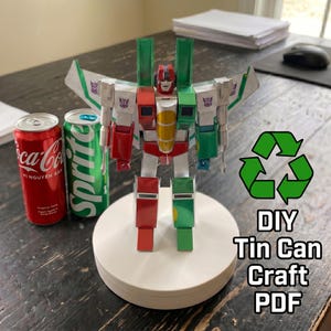 May include: A robot figure constructed from recycled aluminum cans, featuring red, green, and silver panels with purple Decepticon insignias. The figure stands on a white circular base next to a red Coca-Cola can and a green Sprite can, with text reading DIY Tin Can Craft PDF.
