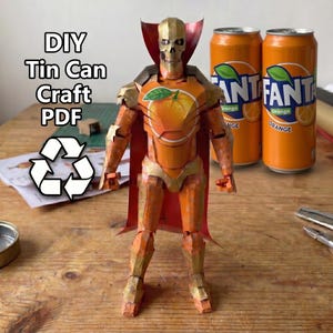 Pode incluir: Uma figura humanoide feita de latas de refrigerante Fanta laranja, com cabeça de caveira, placa peitoral com gráfico de laranja e capa vermelha. O texto diz DIY Tin Can Craft PDF acima de um símbolo de reciclagem, com duas latas ao lado.