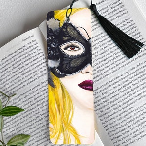 Puede incluir: Un marcapáginas con una ilustración de acuarela de una mujer que lleva una máscara de carnaval negra con detalles dorados. Sus ojos son visibles a través de la máscara y lleva un pintalabios rojo brillante. Su cabello es rubio y fluido.