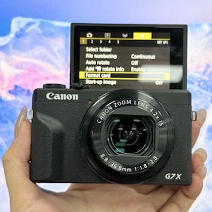 以下が含まれることがあります： フリップアップスクリーンを備えた黒いCanon G7Xデジタルカメラ。画面にはSelect folder、File numbering、Auto rotate、Add rotate info、Format card、Start-up imageなどのオプションを含むメニューが表示されています。レンズにはCanon Zoom Lens 4.2x ISと記載されています。