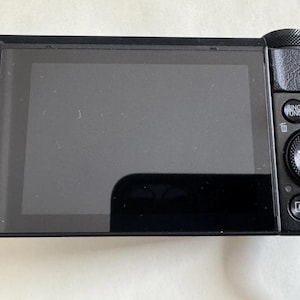 May include: A black digital camera featuring a large rectangular rear display screen. To the right of the screen are several circular control buttons, including a central dial labeled SET and INFO, a red recording button, and a menu button.
