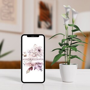 May include: A black smartphone with a white screen displaying a floral design and the text "AVADA KEDAVRA". The phone is sitting on a white table next to a potted plant with white calla lilies.