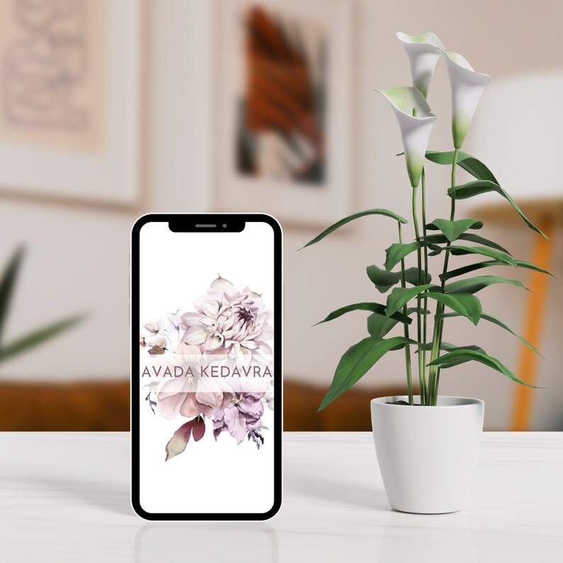 May include: A black smartphone with a white screen displaying a floral design and the text "AVADA KEDAVRA". The phone is sitting on a white table next to a potted plant with white calla lilies.