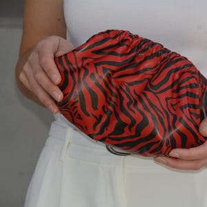 May include: A red and black zebra print clutch bag. The bag is gathered at the top and held by a person. The bag's pattern is a bold, eye-catching design. The background is a neutral colour.