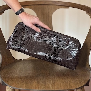 Può includere: Una pochette marrone scuro con una finitura lucida e testurizzata che imita l'aspetto della pelle di coccodrillo. La borsa rettangolare presenta una struttura rigida e una superficie liscia e lucida che riflette la luce sul motivo in rilievo.