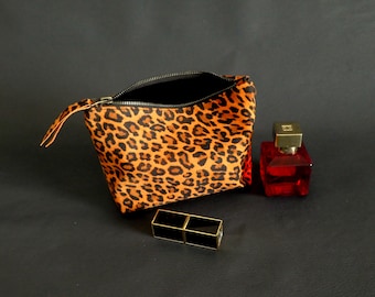 May include: A small leopard print makeup bag with a zipper closure. The bag is open and there is a red perfume bottle and a black lipstick on a black surface.