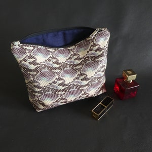May include: A snakeskin-patterned zippered pouch with a blue lining, open to reveal the interior. A small red bottle of perfume and a gold lipstick case are on a black surface.