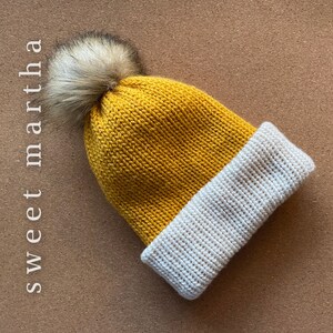 May include: A knitted hat with a white brim and a yellow crown. The hat has a fluffy brown pom pom on top.