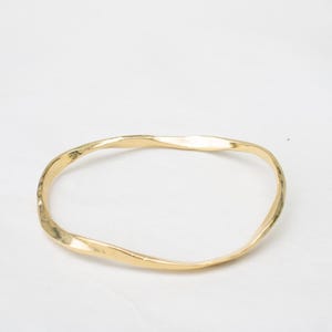 Könnte beinhalten: Ein goldfarbenes Armreif-Armband mit gehämmerten Texturen. Das Armband hat eine unregelmäßige, wellenförmige Form und ist vor einem weißen Hintergrund platziert. Das Schmuckstück ist ein schlichtes, elegantes Accessoire.