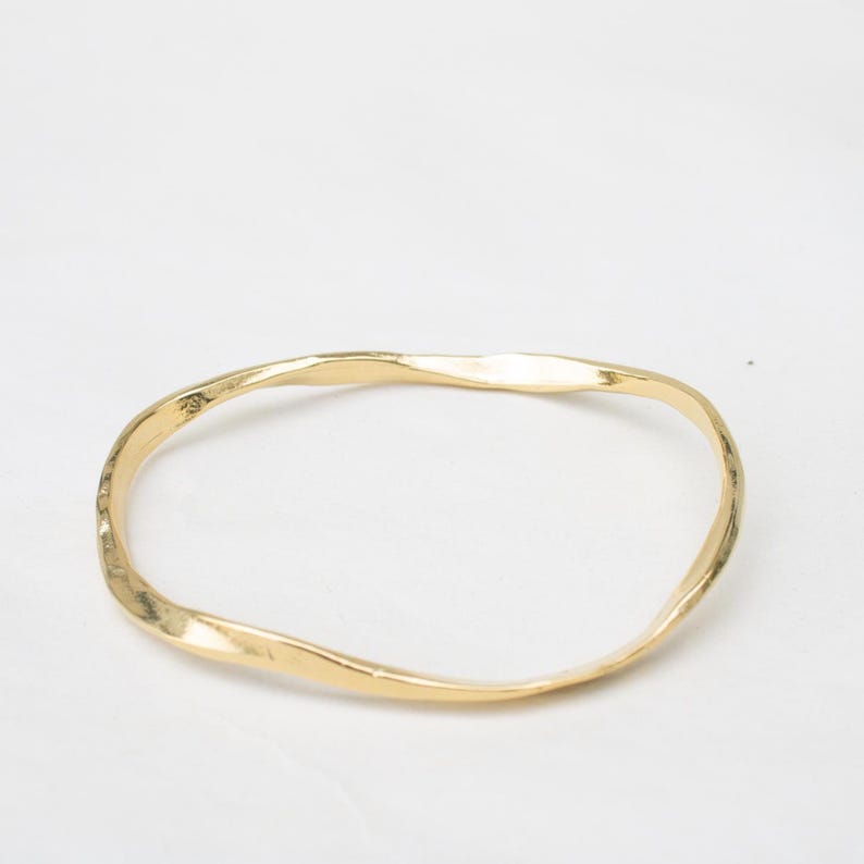 Puede incluir: Un brazalete de color dorado con una textura martillada. El brazalete tiene una forma irregular y ondulada y est&aacute; sobre un fondo blanco. La joya es un accesorio sencillo y elegante.