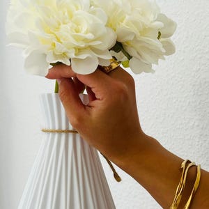 Puede incluir: Un jarr&oacute;n de cer&aacute;mica blanco y acanalado con un detalle de cuerda sostiene un ramo de flores grandes de color crema. Las flores son sostenidas por una mano que lleva un anillo de oro y pulseras doradas. El fondo es una pared blanca lisa.