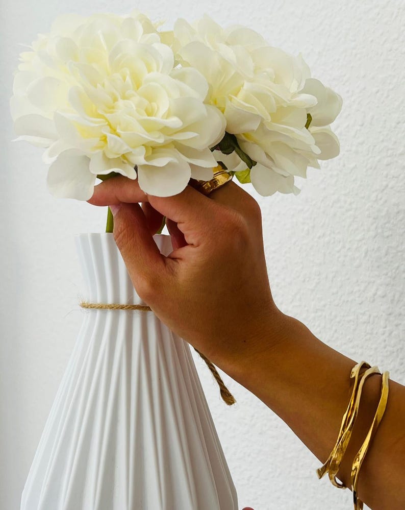 Puede incluir: Un jarr&oacute;n de cer&aacute;mica blanco y acanalado con un detalle de cuerda sostiene un ramo de flores grandes de color crema. Las flores son sostenidas por una mano que lleva un anillo de oro y pulseras doradas. El fondo es una pared blanca lisa.