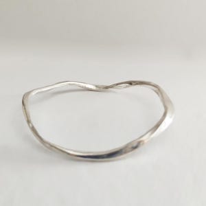 May include: A silver-coloured, wavy bangle bracelet. The bracelet has an organic, irregular shape, with a smooth, reflective surface. The design is minimalist and modern. The bracelet's diameter is approximately 6.35 cm.