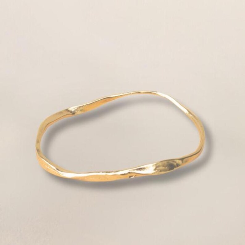 Puede incluir: Un brazalete dorado con un dise&ntilde;o texturizado y org&aacute;nico. El brazalete tiene una forma ligeramente irregular, con un acabado liso y pulido. La joya est&aacute; colocada sobre un fondo neutro.