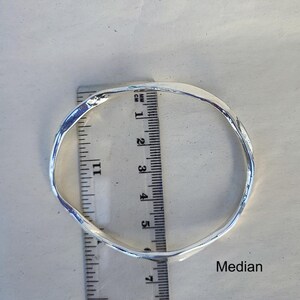 Puede incluir: Un brazalete de metal plateado y retorcido. El brazalete se mide con una regla, mostrando un di&aacute;metro de aproximadamente 6,35 cm. La palabra "Median" est&aacute; impresa en la superficie.