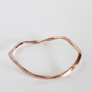 Può includere: Un bracciale a cerchio ondulato color oro rosa. Il bracciale ha una forma organica e irregolare, con una superficie liscia e lucida. Il metallo riflette la luce, creando riflessi e ombre. Il bracciale è posto su uno sfondo bianco.