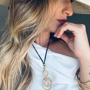 Puede incluir: Collar con colgante dorado con cord&oacute;n negro. El colgante presenta una serie de c&iacute;rculos abiertos interconectados. El collar se lleva con una parte superior blanca y pulseras doradas. Tambi&eacute;n se ve un sombrero de paja con una banda estampada.