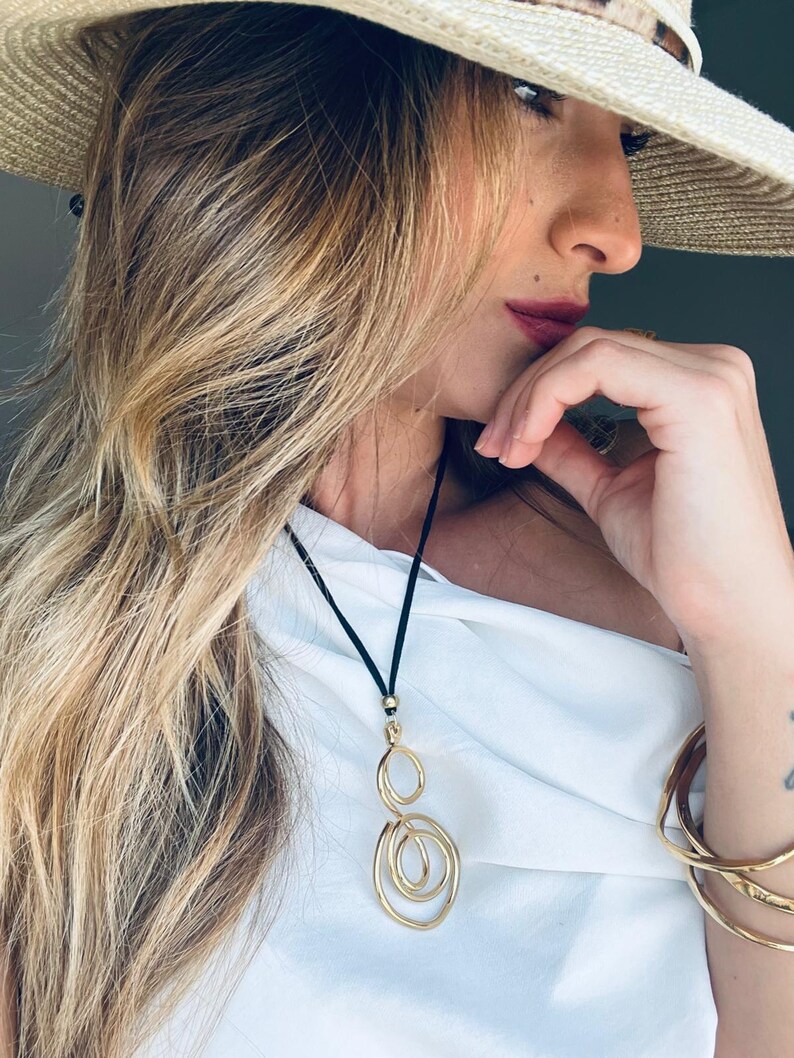 Puede incluir: Collar con colgante dorado con cord&oacute;n negro. El colgante presenta una serie de c&iacute;rculos abiertos interconectados. El collar se lleva con una parte superior blanca y pulseras doradas. Tambi&eacute;n se ve un sombrero de paja con una banda estampada.