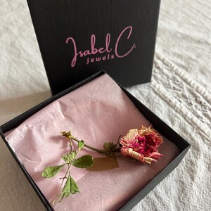 Puede incluir: Una caja de joyer&iacute;a negra con el texto "Isabel C jewels" en escritura rosa. En el interior, una rosa seca con p&eacute;talos rosas y crema descansa sobre papel de seda rosa. La caja est&aacute; abierta, revelando el contenido.