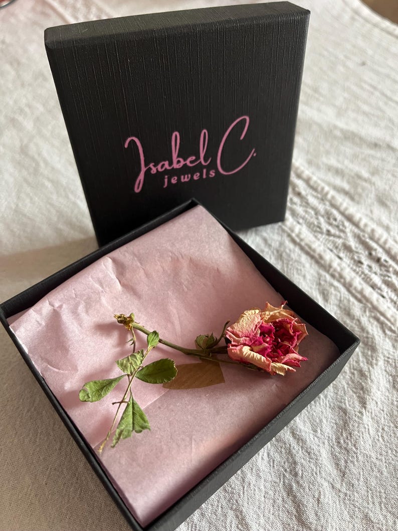 Puede incluir: Una caja de joyer&iacute;a negra con el texto "Isabel C jewels" en escritura rosa. En el interior, una rosa seca con p&eacute;talos rosas y crema descansa sobre papel de seda rosa. La caja est&aacute; abierta, revelando el contenido.